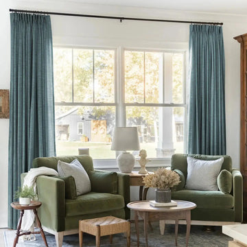 A rustic living room interior featuring two olive green velvet armchairs, striped throw pillows, blue drapes, and a large window overlooking a grey barn with two decorative wreaths and a white table lamp with a bust statue