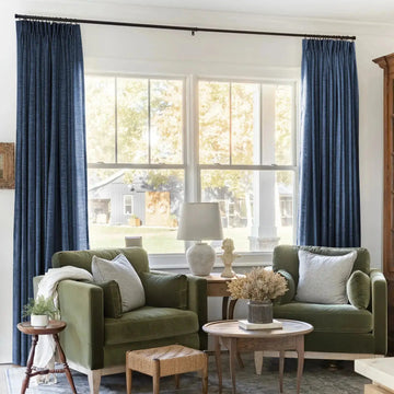 Interior photograph of a cozy living room with two olive green velvet armchairs flanking a large window offering a view of a white barn with wreaths outside, highlighted by dark blue pleated curtains and a decorative bust sculpture