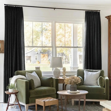 Photograph of a cozy living room interior featuring two plush olive green velvet armchairs with striped pillows, black pleated curtains, and a classical bust overlooking a rural landscape with a dark gray barn and white porch columns