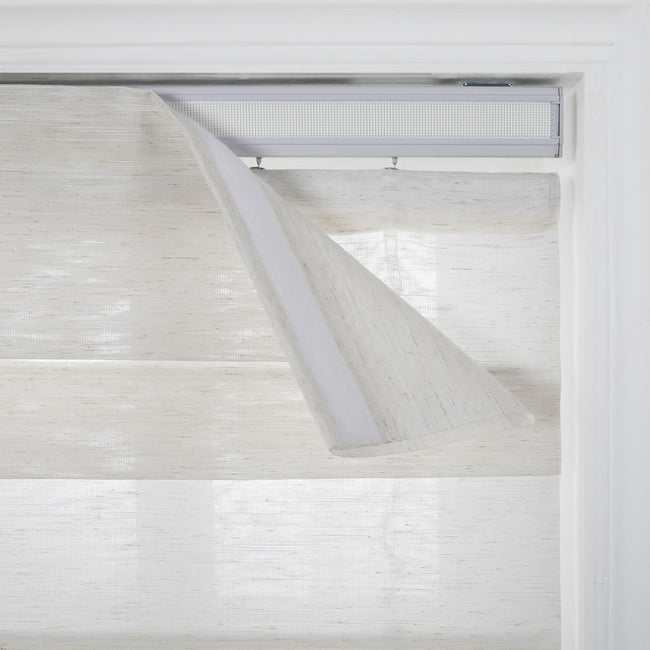 Photography of a dual-layer window blind with natural woven light beige fabric installed in a white window frame, showing the silver headrail and a partially detached valance revealing a hook-and-loop fastener