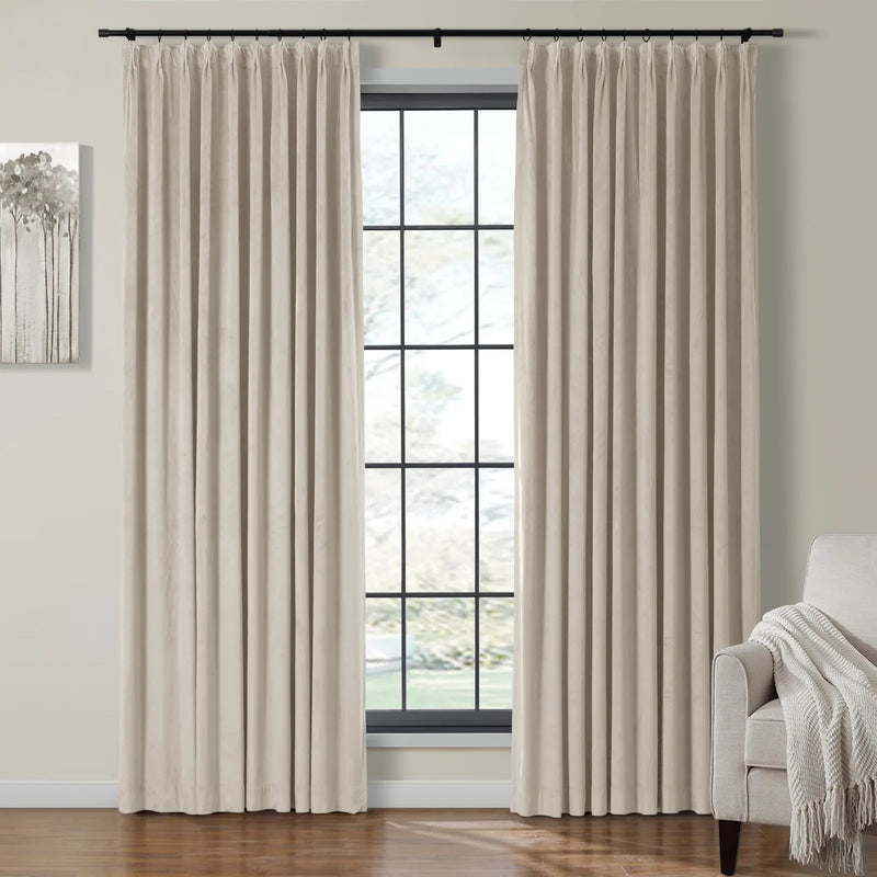 Photograph of an interior room with beige pinch pleat curtains on a black rod framing a large grid window overlooking a green landscape, featuring abstract grey tree wall art and a neutral armchair with a white fringed throw