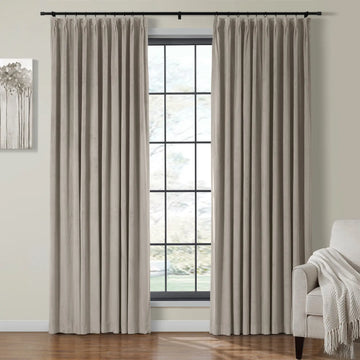 Elegant beige velvet pleated drapes on a black rod adorning a large window with black grid panes revealing a soft green outdoor view, alongside a grey armchair with a white knit throw and an abstract tree artwork in a contemporary interior