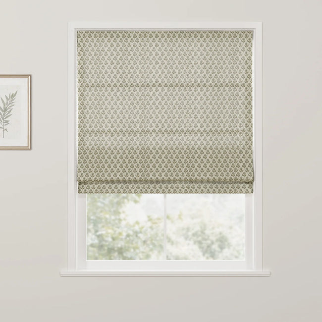 Photograph of an interior window featuring a partially lowered cream Roman blind with a repeating olive green floral pattern, offering a blurred view of green foliage, complemented by a framed botanical print on the white wall