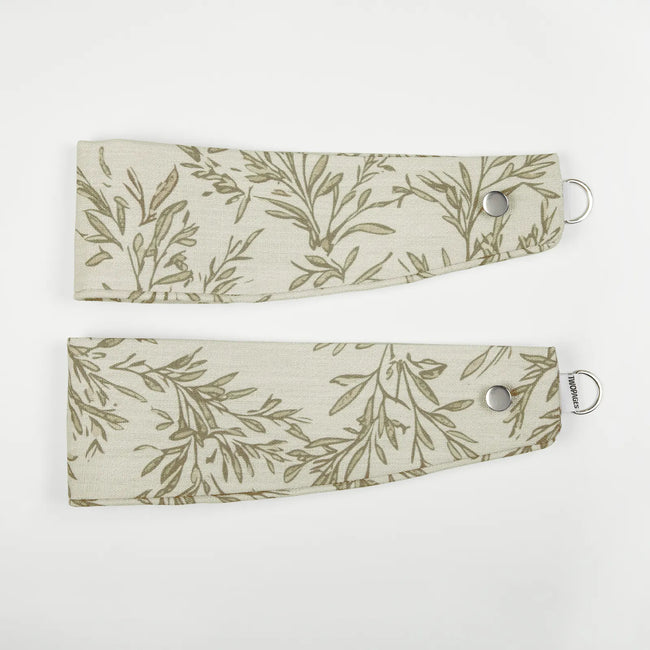 Two light beige fabric curtain tie-backs featuring a green olive branch pattern, each with a silver snap button and D-ring, one displaying a TWOPAGES label, laid flat on a white background