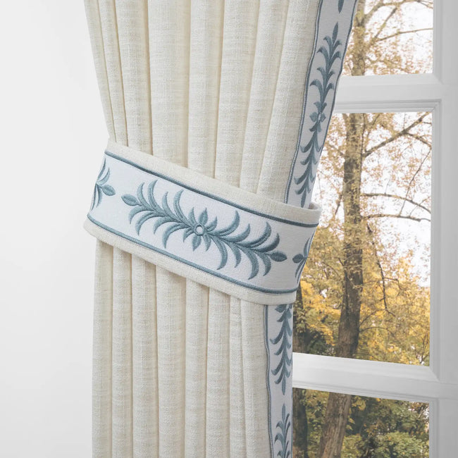 A cream linen curtain tied back with an elaborate embroidered blue-green floral sash revealing autumn trees with yellow leaves through a white paneled window