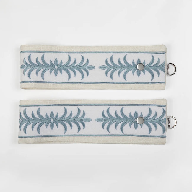 Two TWOPAGES fabric tiebacks with pale blue acanthus embroidery on a white stripe, flanked by natural beige borders, featuring silver snap closures and D-rings displayed on a white background