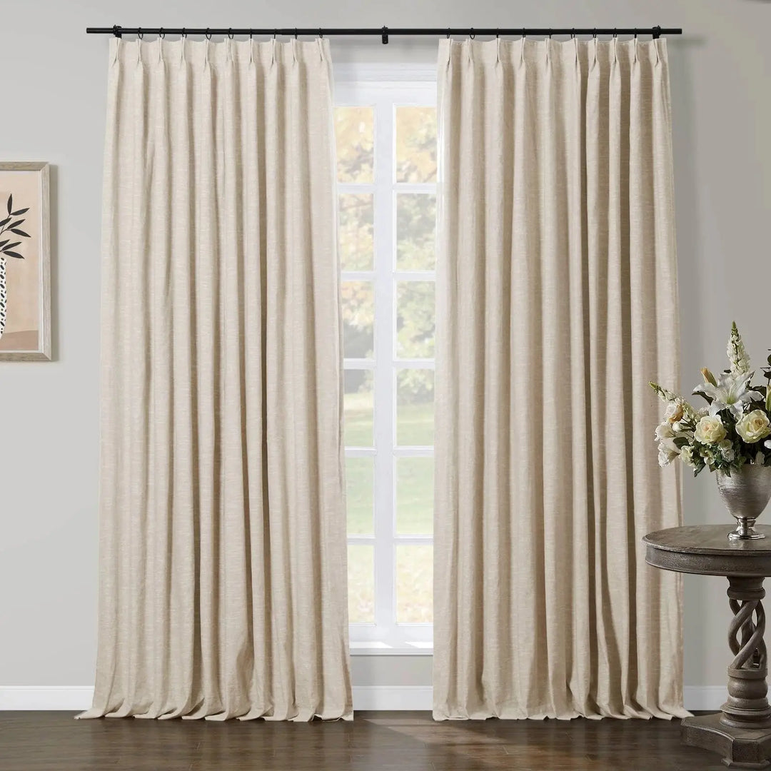 Light beige pleated curtains on a black rod frame a large window with a soft green outdoor view in an elegant room, featuring a twisted dark wood side table with a silver vase of white and cream flowers