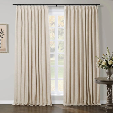 Light beige pleated curtains on a black rod frame a large window with a soft green outdoor view in an elegant room, featuring a twisted dark wood side table with a silver vase of white and cream flowers