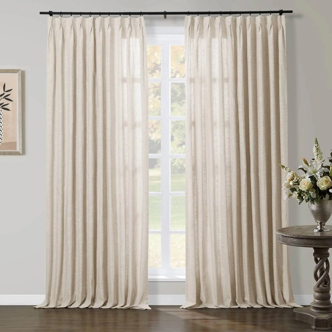 A bright interior features floor-length beige linen-look pinch pleat curtains on a black rod framing a large white grid window with an outdoor view of lush green foliage, complemented by a dark wood twisted pedestal table holding a silver vase of white and cream flowers