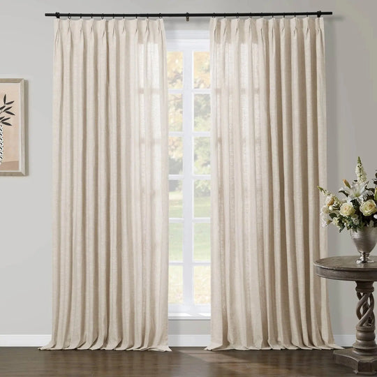 A bright interior features floor-length beige linen-look pinch pleat curtains on a black rod framing a large white grid window with an outdoor view of lush green foliage, complemented by a dark wood twisted pedestal table holding a silver vase of white and cream flowers