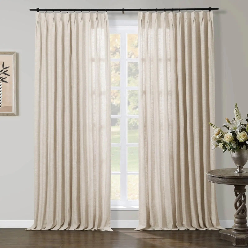 A bright interior features floor-length beige linen-look pinch pleat curtains on a black rod framing a large white grid window with an outdoor view of lush green foliage, complemented by a dark wood twisted pedestal table holding a silver vase of white and cream flowers