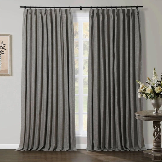 Grey pleated curtains hang from a black rod over a window, flanked by abstract wall art and a silver vase with a white floral arrangement on a carved wooden table
