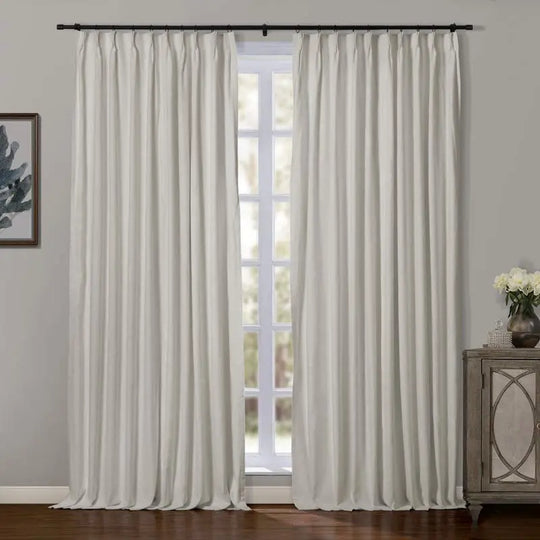 Photograph of cream-colored pinch-pleat blackout curtains on a black rod flanking a bright window revealing outdoor greenery, with a decorative wooden cabinet and botanical framed art contributing to a serene room aesthetic