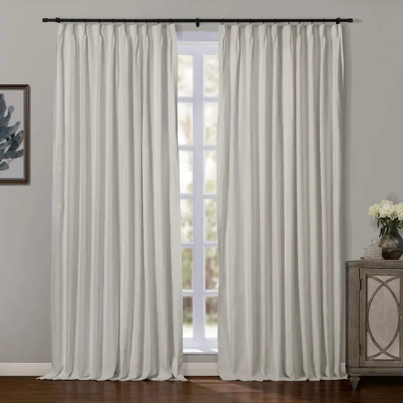 Photograph of cream-colored pinch-pleat blackout curtains on a black rod flanking a bright window revealing outdoor greenery, with a decorative wooden cabinet and botanical framed art contributing to a serene room aesthetic