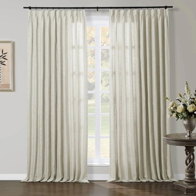 A pair of neutral cream linen blend pinch pleat curtains with a black rod hangs over a multi-pane window offering a lush outdoor view, accented by a white and yellow floral arrangement in a silver vase on a decorative carved wooden side table