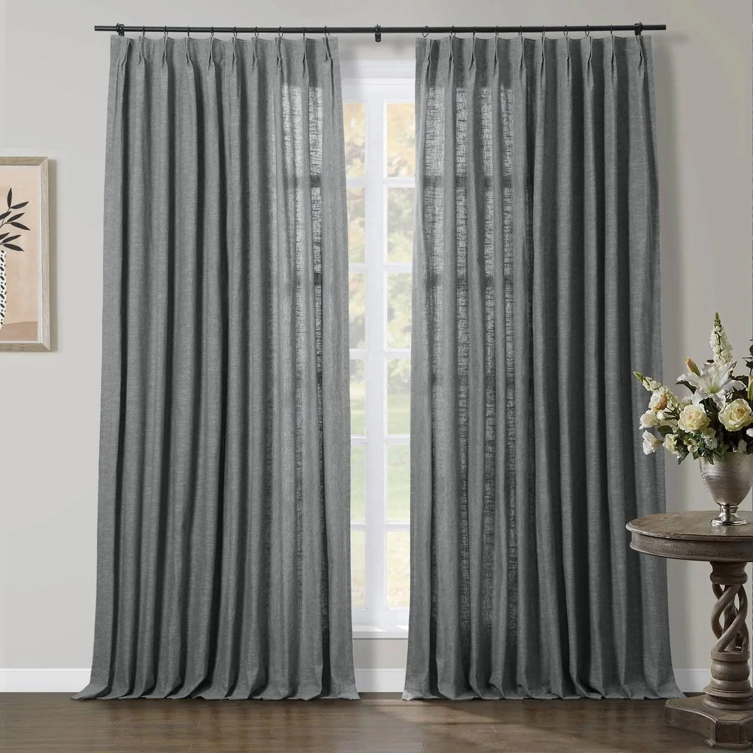 A room featuring a large window dressed with grey linen blend pinch pleat curtains on a dark rod, a decorative carved wooden side table holding a vase of white and cream flowers, and a framed abstract plant artwork on the left wall
