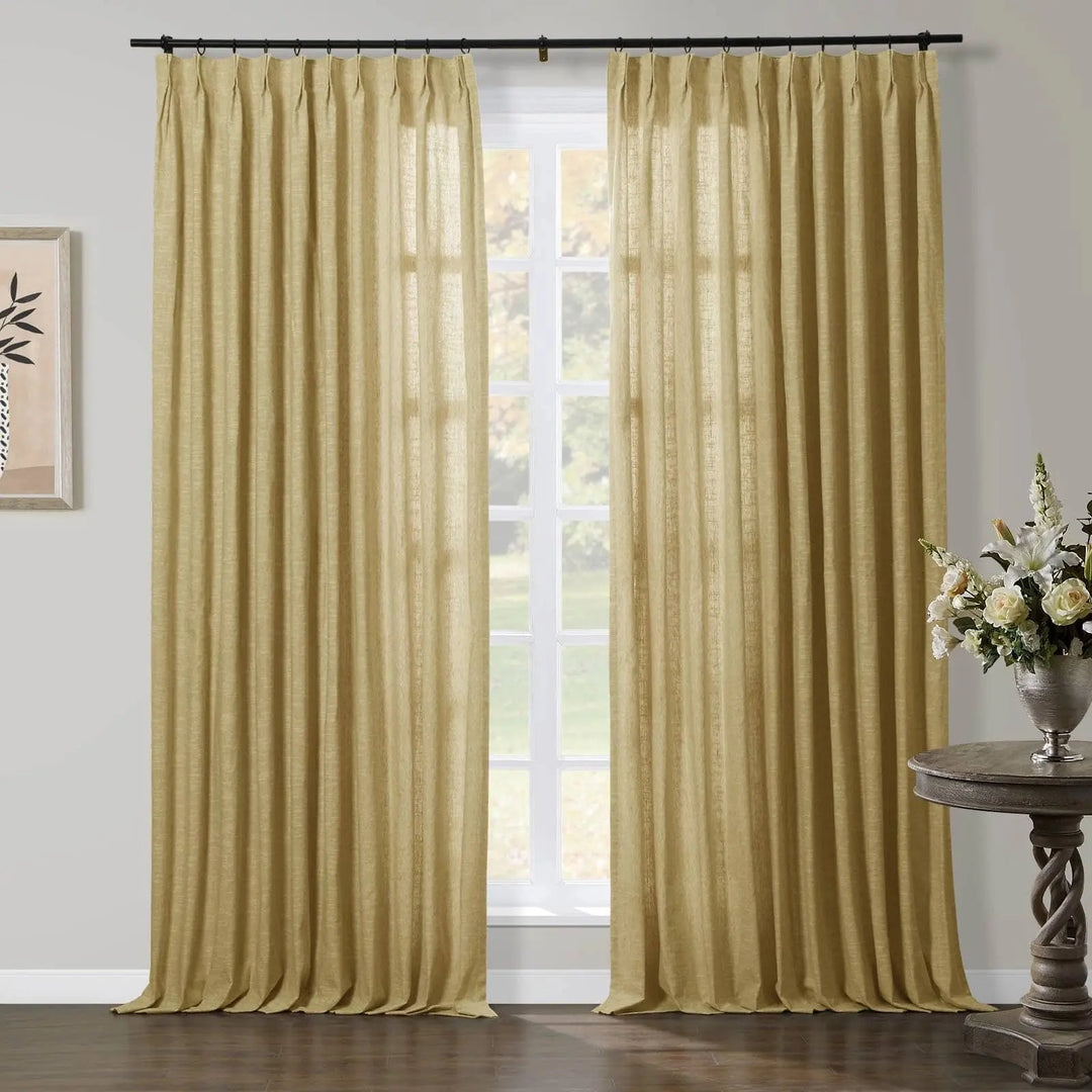 A pair of pleated, textured gold curtains framing a sunlit window in a living room, accompanied by a rustic carved wood side table adorned with white roses and lilies in a silver vase, and a framed abstract artwork on the left wall