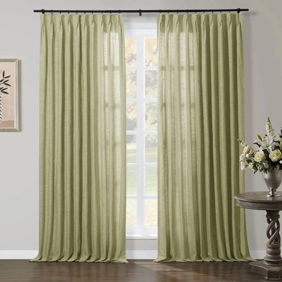 A pair of light olive-green linen pinch pleat curtains hanging from a black rod in a room with a window overlooking trees, featuring an ornate wooden side table with a silver vase holding white and cream flowers and framed abstract plant art