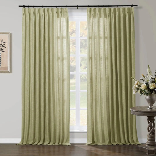 A pair of light olive-green linen pinch pleat curtains hanging from a black rod in a room with a window overlooking trees, featuring an ornate wooden side table with a silver vase holding white and cream flowers and framed abstract plant art