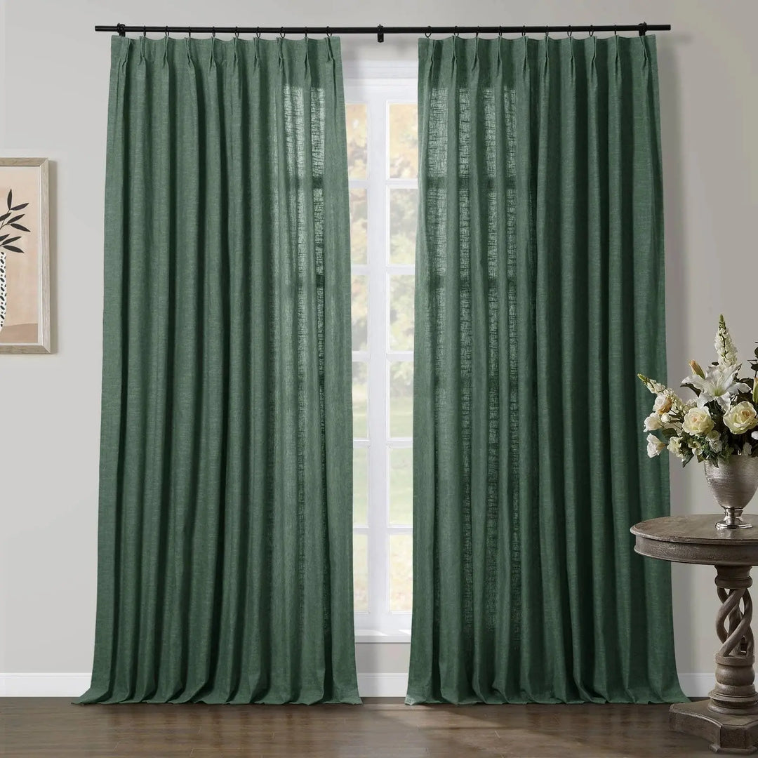 A pair of textured forest green pinch pleat curtains covering a window in an interior room with a white and cream floral arrangement in a silver vase on a dark carved wood side table