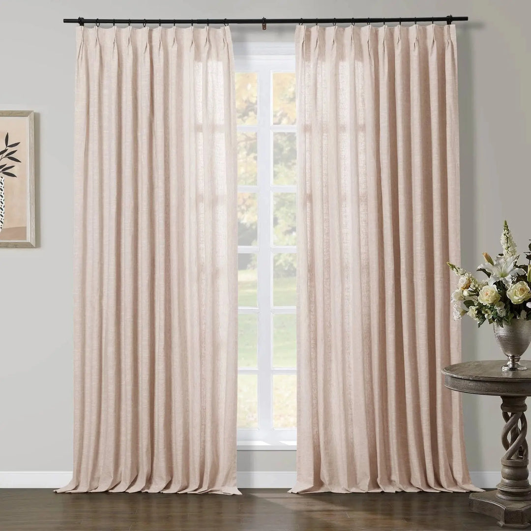 Pleated beige linen curtains softly illuminate a room through a window overlooking green trees, accented by a rustic carved console table with white and yellow flowers in a silver vase, and minimalist framed art