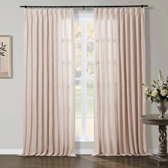 Pleated beige linen curtains softly illuminate a room through a window overlooking green trees, accented by a rustic carved console table with white and yellow flowers in a silver vase, and minimalist framed art