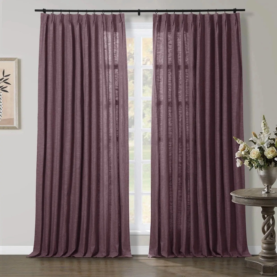 Deep plum pinch pleat curtains with a linen texture hanging floor-length over a window, with a framed botanical print on the left and a white floral arrangement on a carved wooden side table to the right