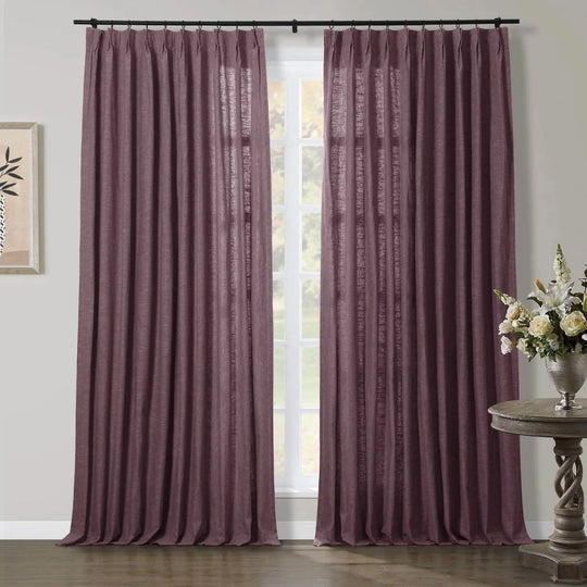 Deep plum pinch pleat curtains with a linen texture hanging floor-length over a window, with a framed botanical print on the left and a white floral arrangement on a carved wooden side table to the right