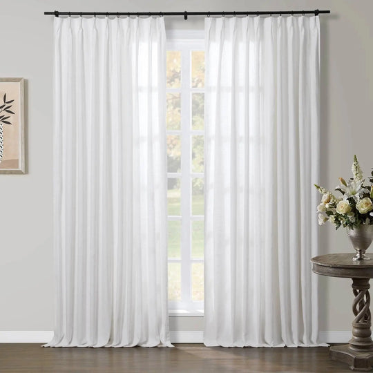 Photograph of sheer white pinch-pleat curtains on a black rod in a bright room with a multi-pane window overlooking a lush green outdoor view, featuring a framed botanical print and an ornate wooden side table holding a silver vase of white and cream flowers