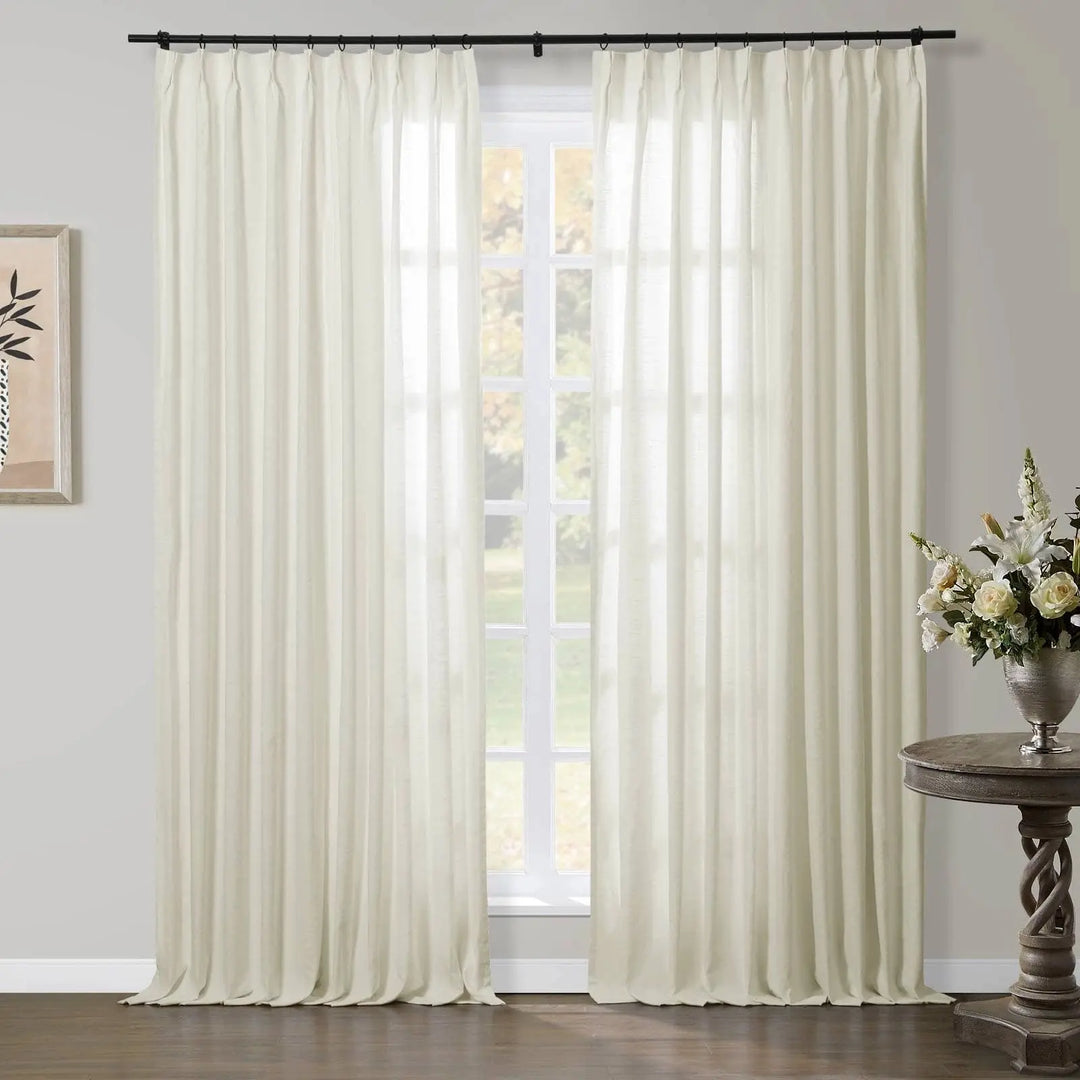 Creamy white pinch-pleat sheer curtains on a black rod in a bright room, beside a distressed wood table with a silver vase of white flowers and framed minimalist botanical art