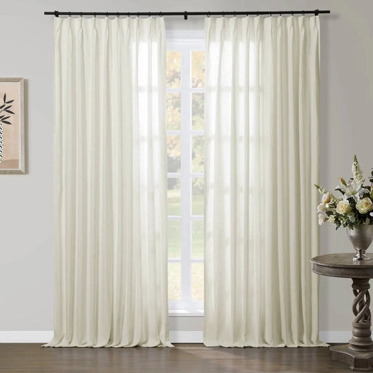Creamy white pinch-pleat sheer curtains on a black rod in a bright room, beside a distressed wood table with a silver vase of white flowers and framed minimalist botanical art