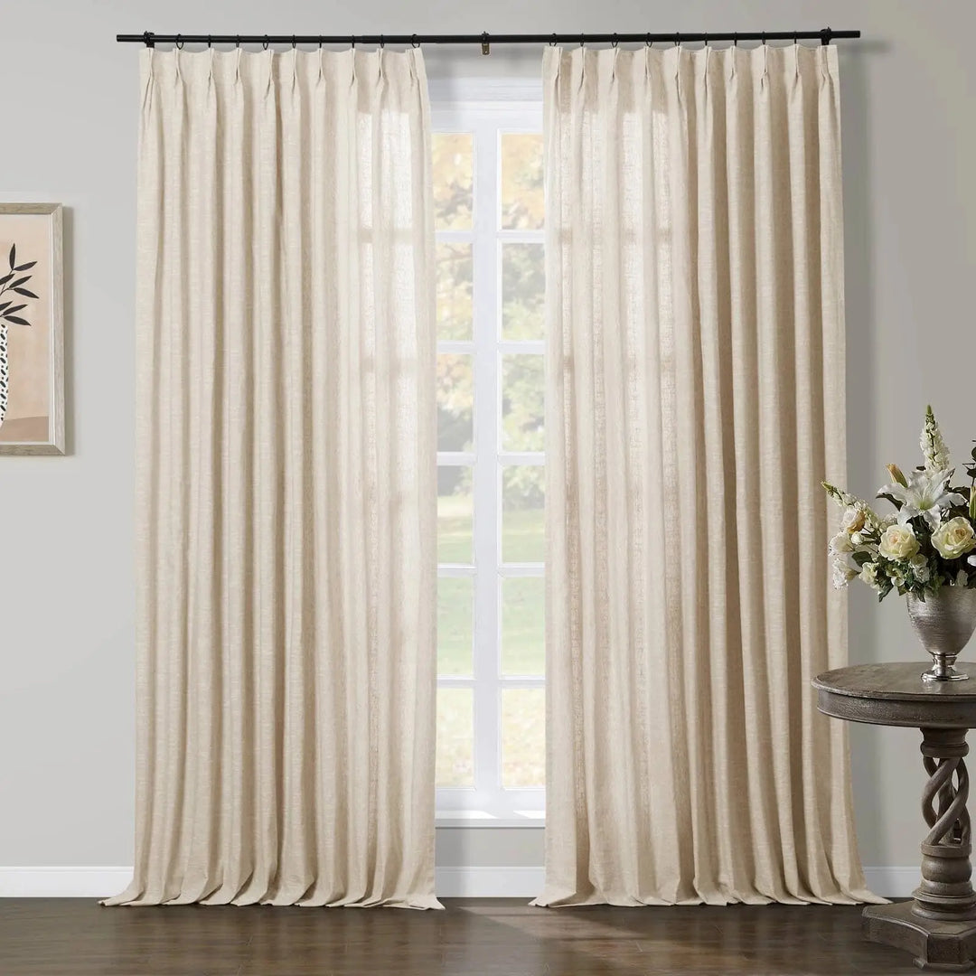 Natural linen beige pleated curtains on a black rod frame a multipane window looking out to outdoor greenery, with a dark wood floor, an ornate carved side table holding white flowers in a silver vase, and minimalist framed art