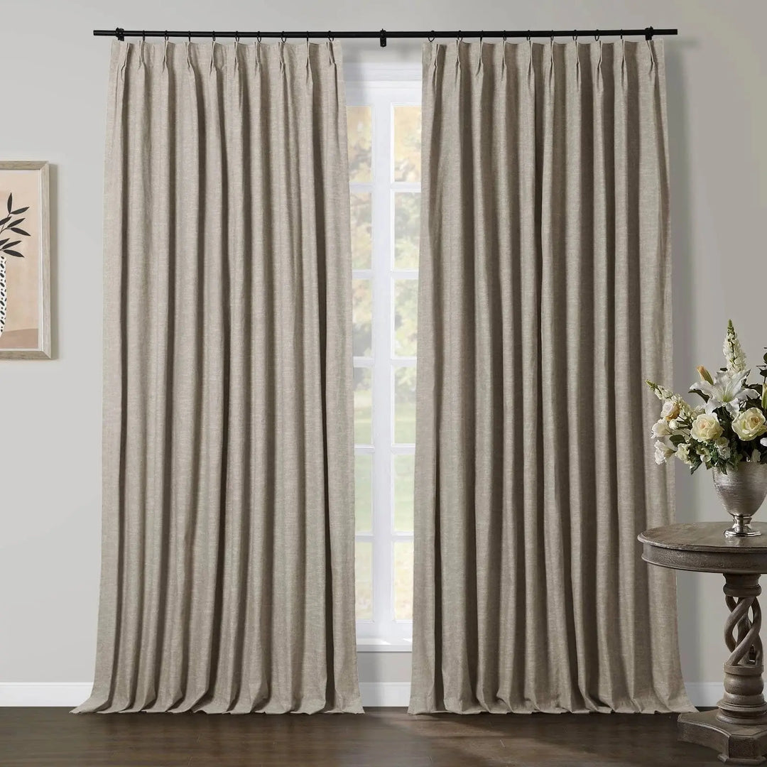 Floor-length natural linen pleated drapery panels on a black rod frame a white gridded window in a modern room with a dark wood floor and a decorative side table holding a white floral arrangement
