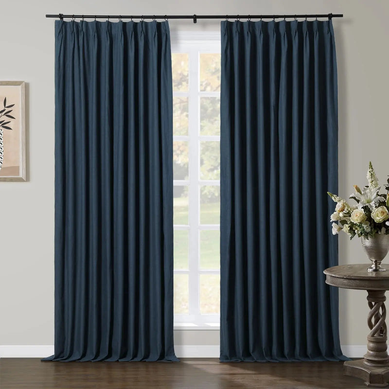 Photography of an interior room featuring elegant dark blue pinch-pleat curtains framing a window that looks out onto a green outdoor landscape, with a decorative wooden console table holding a silver vase of white and yellow flowers