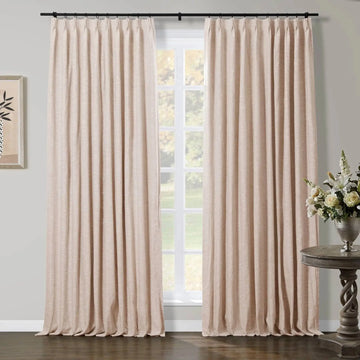 Natural beige linen-look pleated curtains on a black rod in a room with a white window overlooking a green garden, accented by framed botanical art and an ornate wooden side table with a silver vase of white flowers