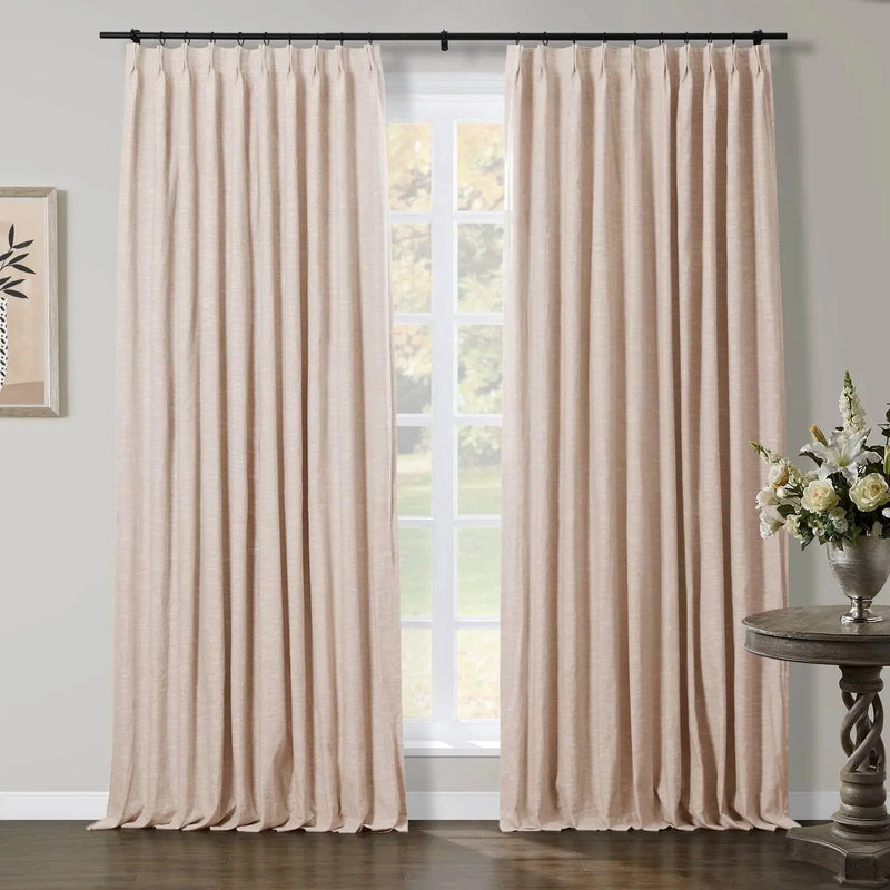 Natural beige linen-look pleated curtains on a black rod in a room with a white window overlooking a green garden, accented by framed botanical art and an ornate wooden side table with a silver vase of white flowers