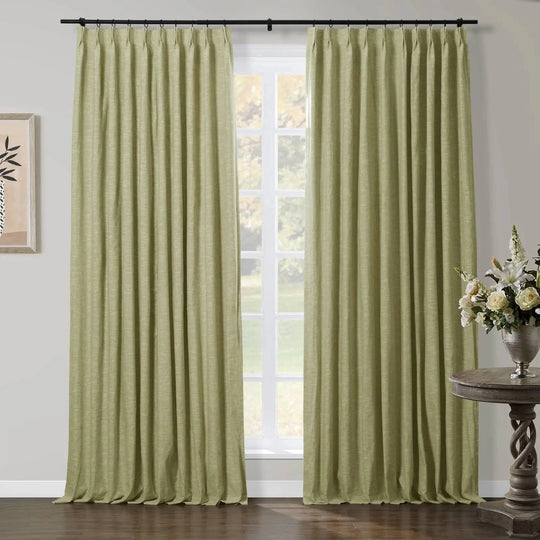 Light green linen-textured curtains hung on a black rod framing a window in a room, with a twisted wooden accent table holding a silver vase of white lilies and cream roses